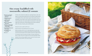 The Picnic round loaf from Simon & Schuster, filled with mozzarella, salami, and tomato, sits invitingly on a plate next to a knife and picnic basket on lush green grass—making it a perfect choice for summer picnics and al fresco dining.
