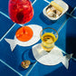 Two Piecework Sardine Cocktail Napkins on a blue tiled surface, each holding a drink, next to an open tin of sardines.