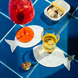 Two Piecework Sardine Cocktail Napkins on a blue tiled surface, each holding a drink, next to an open tin of sardines.