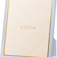 The Aerin Piero Leather Frame Collection features a rectangular frame with a white leather border and gold detailing, standing upright for an elegant display on any flat surface. The brand name "AERIN" is displayed in the center.