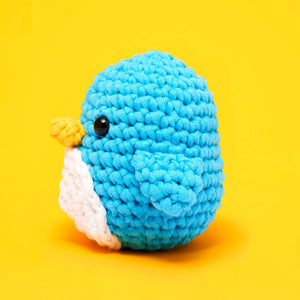 Pierre the Penguin Beginner Crochet Kit by The Woobles features a crocheted blue penguin with a white belly and yellow beak, set against a bright yellow background—perfect for beginner crochet enthusiasts or fans of The Woobles’ charming style.