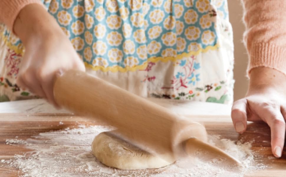 Person rolling out dough with a wooden rolling pin on a floured surface, dressed in a patterned apron and peach-colored sweater—just as taught by Kate Lebo in Pie School from Penguin Random House, mastering techniques for irresistibly delicious pies.