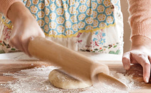 Person rolling out dough with a wooden rolling pin on a floured surface, dressed in a patterned apron and peach-colored sweater—just as taught by Kate Lebo in Pie School from Penguin Random House, mastering techniques for irresistibly delicious pies.