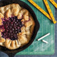 A rustic berry galette inspired by pie recipes from Penguin Random House’s Pie School sits in a cast iron skillet on a green cutting mat, with chalk and folded rulers nearby.