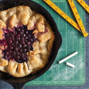 A rustic berry galette inspired by pie recipes from Penguin Random House’s Pie School sits in a cast iron skillet on a green cutting mat, with chalk and folded rulers nearby.