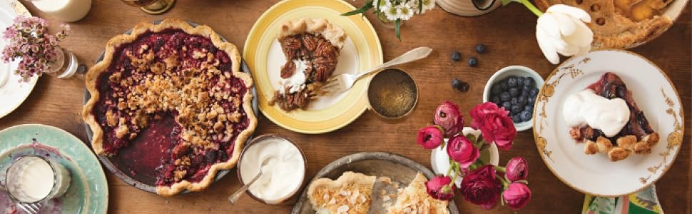 A wooden table adorned with an array of pies, plates holding pie slices, bowls of whipped cream, blueberries, flowers, and gold utensils arranged for serving—an inviting scene reminiscent of the mouthwatering creations featured in Pie School by Penguin Random House, perfect for anyone looking to discover inspiring pie recipes.