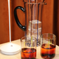A glass pitcher with an ice block, two glasses filled with a red liquid, and a Zafferano Pina Lamp from Zafferano America on a beige surface create an elegant display in front of a brown curtain.