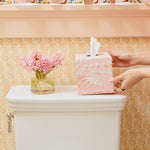 A person sets a pink hand-marbled tissue box from the Marble Bath Collection by Weezie Towels on a white toilet tank beside a glass vase of pink flowers, all against a patterned yellow wall.