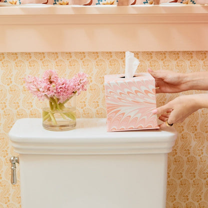 A person sets a pink hand-marbled tissue box from the Marble Bath Collection by Weezie Towels on a white toilet tank beside a glass vase of pink flowers, all against a patterned yellow wall.