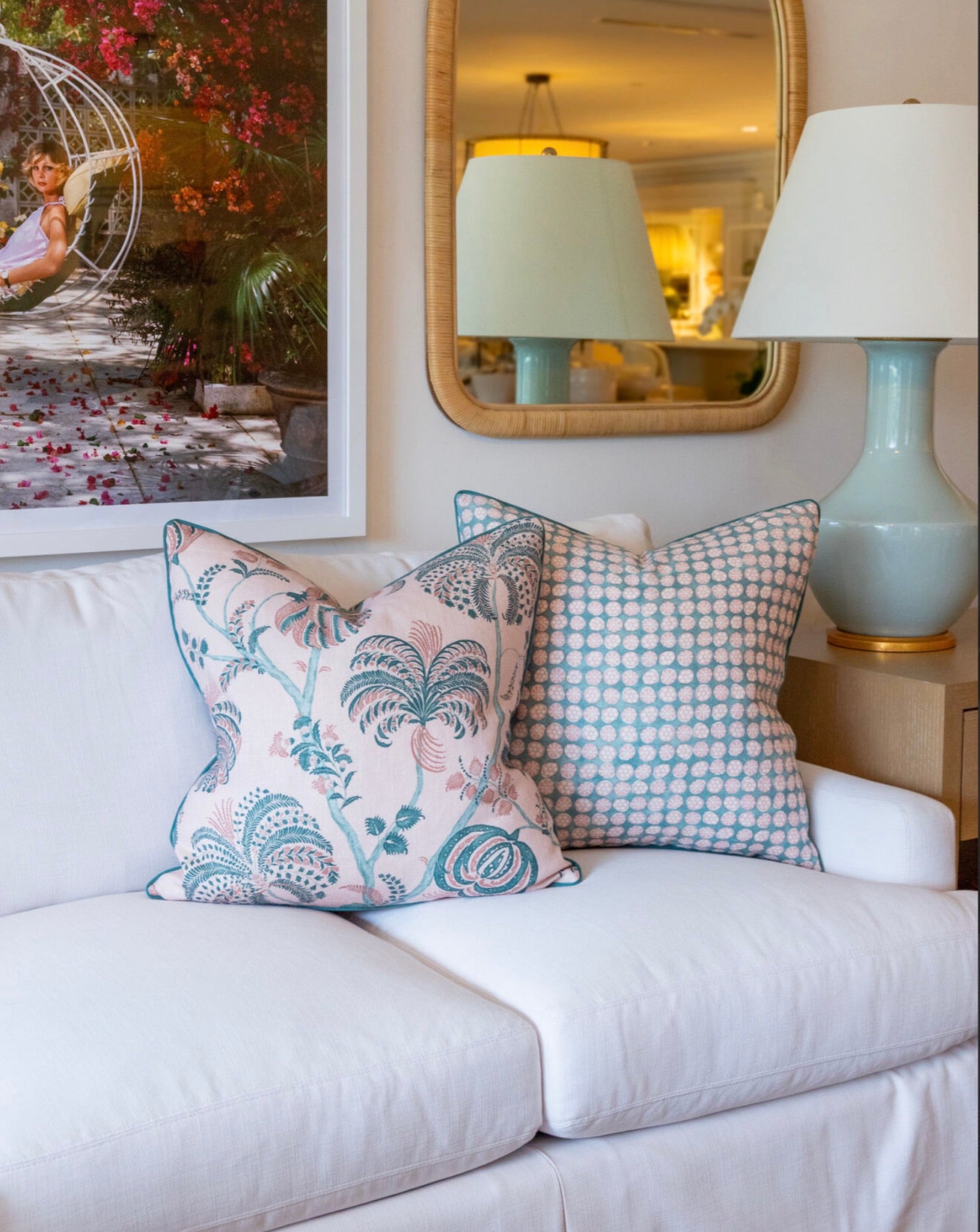 A white sofa with two Boheme Pink and Celadon pillows by Hive Custom Pillows, both down filled for extra comfort, sits next to a lamp beneath a framed photo and a wall mirror.