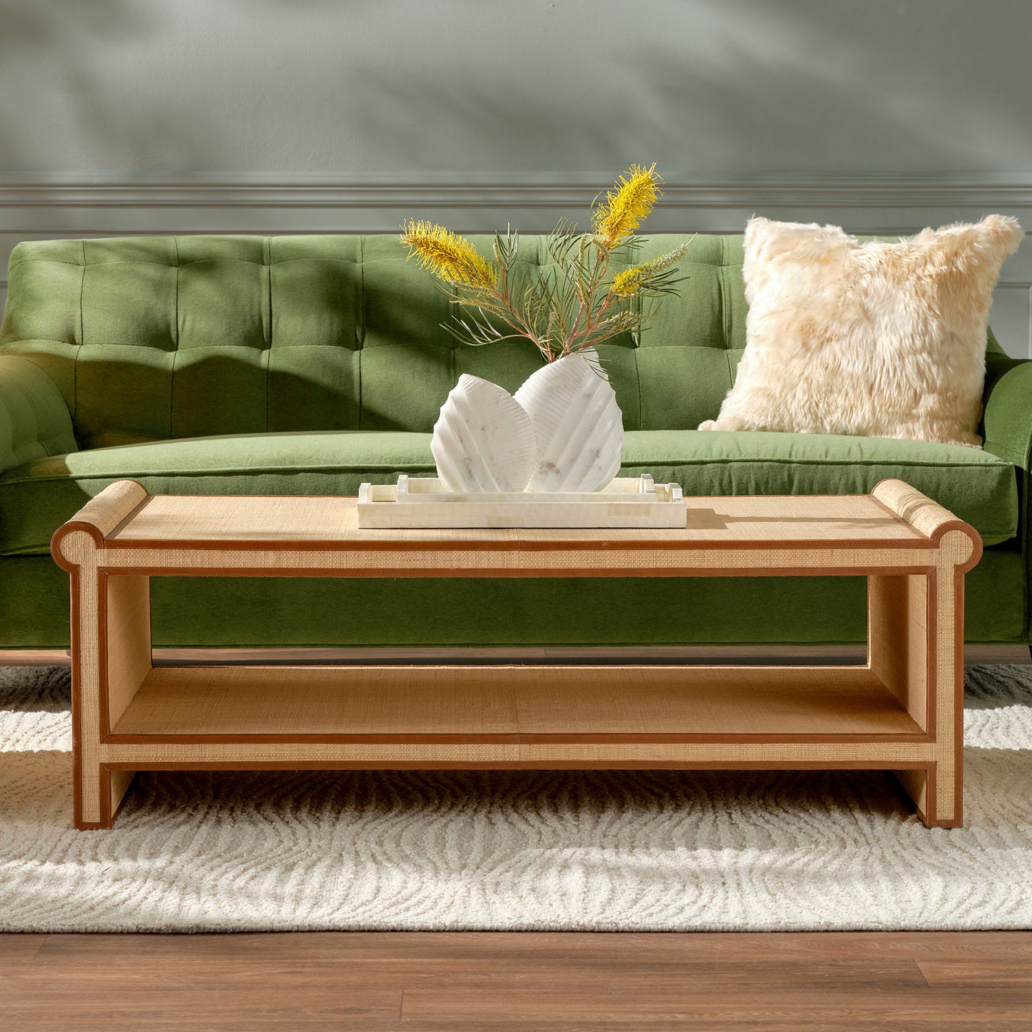 The Matthew Coffee Table by Made Goods, featuring a unique light wood design and a decorative vase, is placed on a cream rug in front of a green sofa with a beige fur pillow.