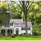 A Place To Call Home by Rizzoli captures the timeless charm of a white two-story house with a stone chimney, front porch, and large windows, all surrounded by trees and a neatly manicured lawn—a beautiful testament to visionary design and architectural excellence.