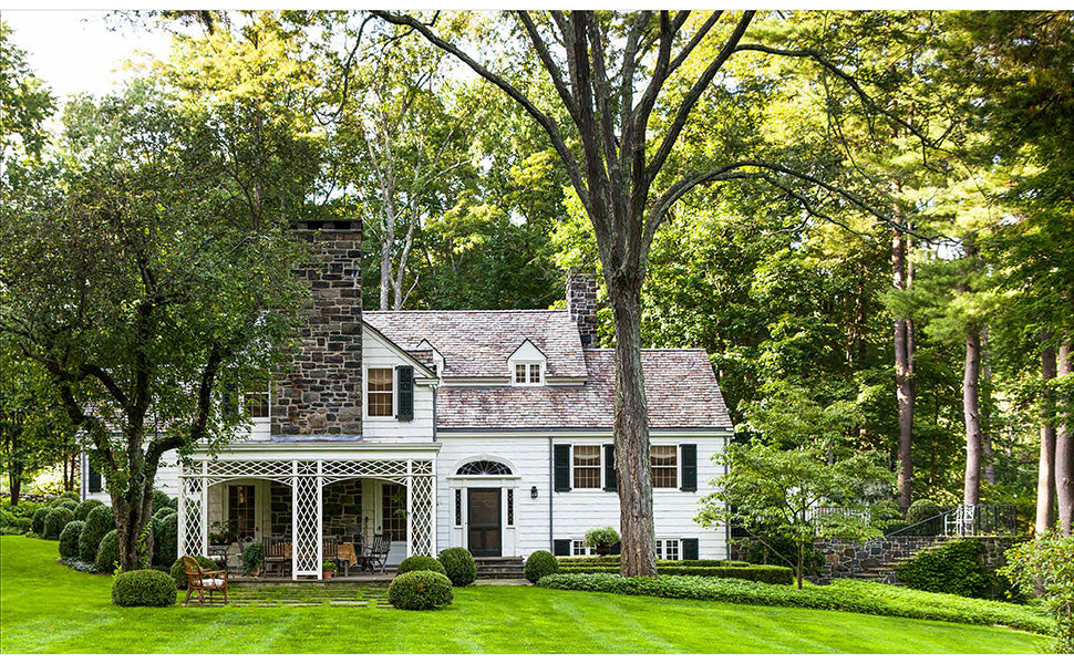A Place To Call Home by Rizzoli captures the timeless charm of a white two-story house with a stone chimney, front porch, and large windows, all surrounded by trees and a neatly manicured lawn—a beautiful testament to visionary design and architectural excellence.