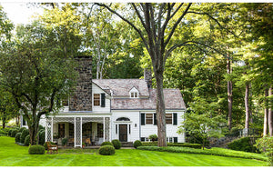 A Place To Call Home by Rizzoli captures the timeless charm of a white two-story house with a stone chimney, front porch, and large windows, all surrounded by trees and a neatly manicured lawn—a beautiful testament to visionary design and architectural excellence.