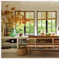 In front of a window dressed with three shades, “A Place To Call Home” by Rizzoli rests on the shelves beneath a classic wooden bench, accompanied by decorative vases, a globe, and a basket. Tree branches arranged in a vase accent the far left, creating an inviting and timeless scene.