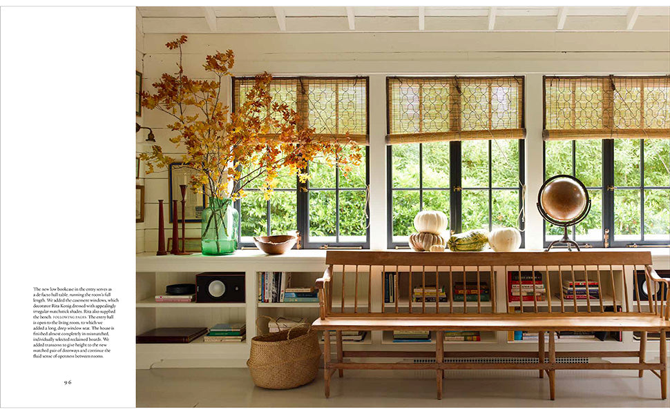 In front of a window dressed with three shades, “A Place To Call Home” by Rizzoli rests on the shelves beneath a classic wooden bench, accompanied by decorative vases, a globe, and a basket. Tree branches arranged in a vase accent the far left, creating an inviting and timeless scene.