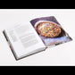 An open copy of Plenty More, Vibrant Vegetable Cooking by Penguin Random House, inspired by Yotam Ottolenghi’s signature style, displays a recipe with text on the left page and a photo of a mouthwatering baked dish topped with circular slices on the right—an ideal choice for fans of creative vegetarian cooking.