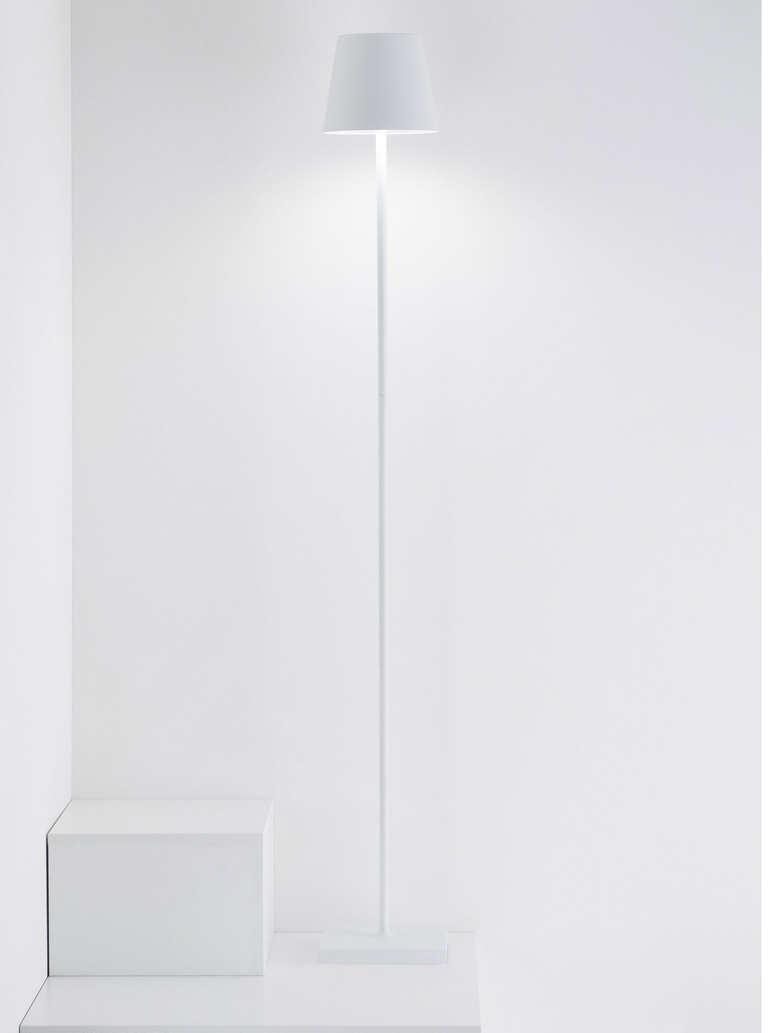 The Zafferano America Poldina Pro L Floor Lamp stands on a white floor by two white cubes against a plain wall, offering cordless convenience and touch dimming.