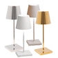 The Poldina Pro Mini Table Lamp Collection by Zafferano America features four modern, cordless lamps with cone shades and square bases in silver, white, beige, and gold—suitable for indoor/outdoor use—displayed on a plain white background.