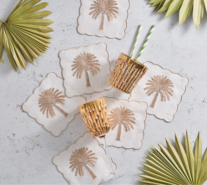 The Kim Seybert Date Palm Cocktail Napkin Set, paired with white coasters, wicker cups with striped straws, and decorative palm leaves on a light grey surface—ideal for breezy gatherings or a standout hostess gift.