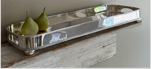 Two green pears sit on the Hôtel Silver Gallery Tray with Bun Feet & Cut-Out Handles, displayed on a rustic wooden shelf against a neutral wall.