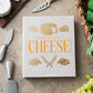 A PrintWorks Cheese Tool Set box sits on a counter surrounded by stainless steel cheese utensils, a cheese board, green napkins, and grapes.