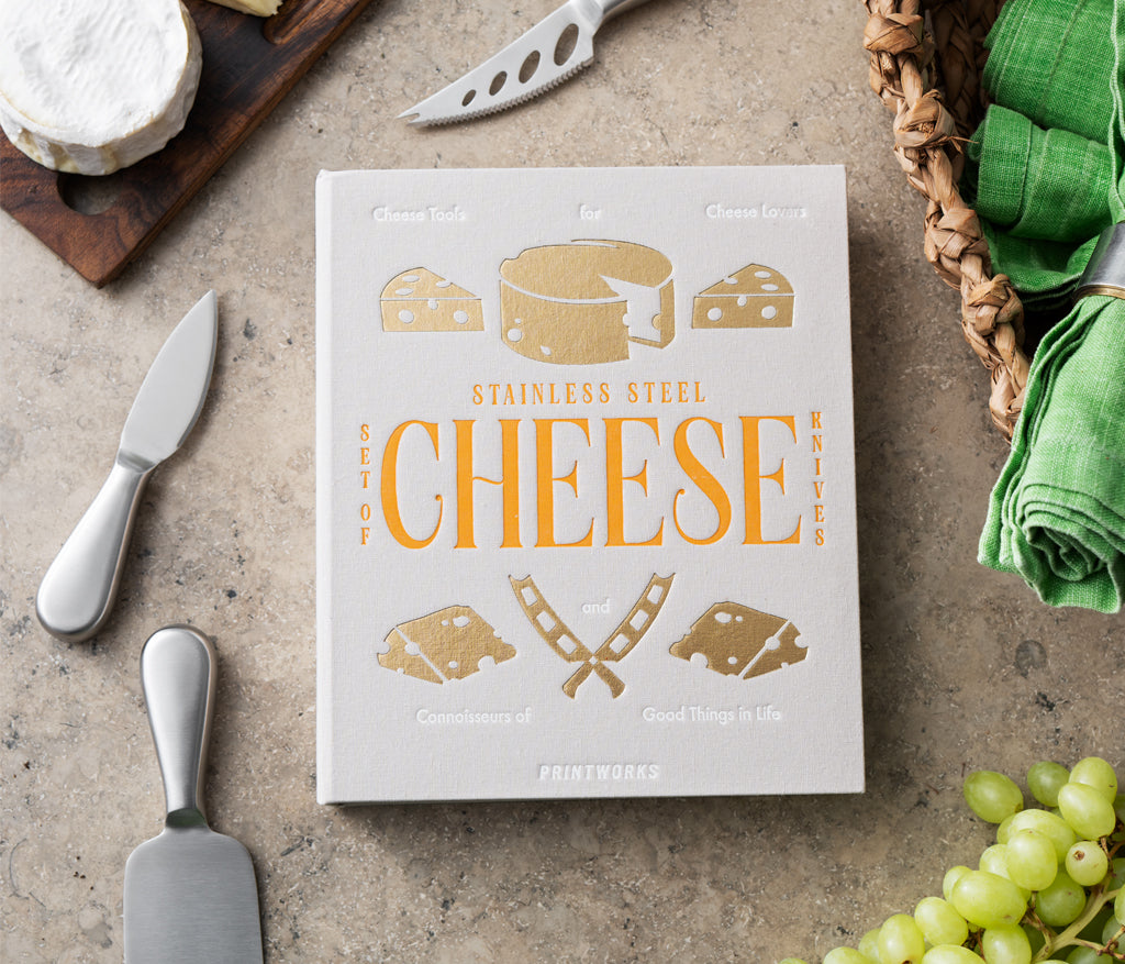 A PrintWorks Cheese Tool Set box sits on a counter surrounded by stainless steel cheese utensils, a cheese board, green napkins, and grapes.