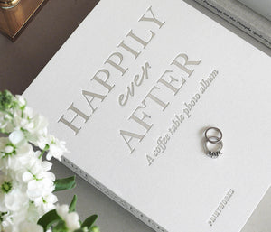 The PrintWorks Happily Ever After Photo Album, in white, rests on a table with two rings and delicate white flowers—perfect for cherishing lasting Wedding Day Memories.