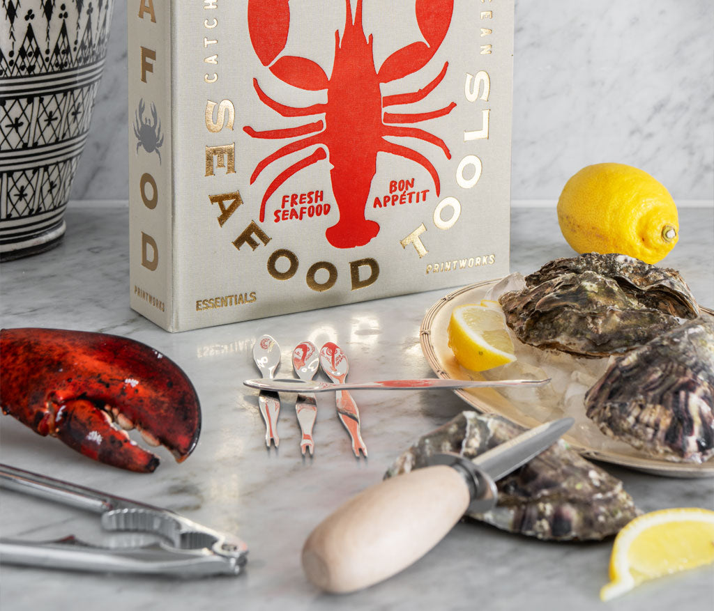 The PrintWorks Seafood Tool Set includes oysters, an oyster knife, shell cracker, lobster claw tool, lemon wedges, and comes in a box labeled "Seafood Tools," all arranged elegantly on a marble surface.