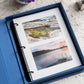 The PrintWorks Moments That Matter The Most Photo Album features an open blue cotton cover coffee table design displaying two landscape photos—rocky terrain with vegetation and a sunset lake dock—on high-quality photo paper.