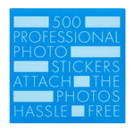 Blue square label with white text: "500 professional acid-free PrintWorks Photo Stickers attach photos hassle free," with several rectangular blanks—made with environmental protection glue—scattered throughout.