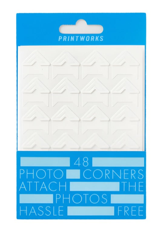 PrintWorks Photo Stickers: Pack of 48 acid-free, matte white, stick-on photo corners with eco-friendly adhesive, neatly arranged in rows and packaged in a blue and white cardboard box.