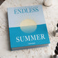 The "Endless Summer Photo Album" by PrintWorks, featuring a sun illustration and a turquoise cloth cover, is displayed on a marble table beside a silver candlestick.