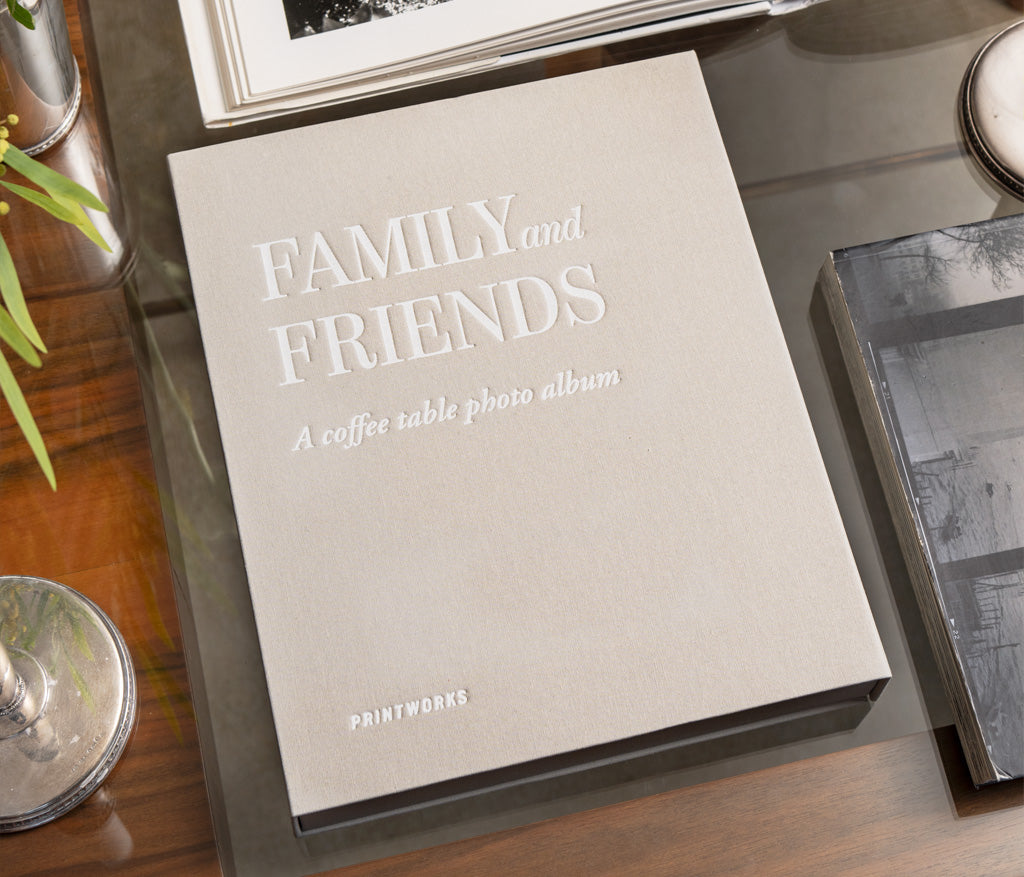 The PrintWorks Family and Friends Photo Album in beige, featuring premium black photo paper, rests on a glass table beside a plant and books—offering chic, organized storage for your treasured memories.