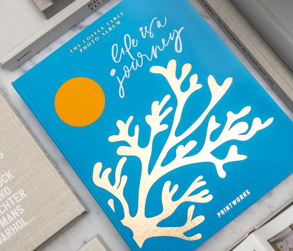 The PrintWorks Life Is A Journey Photo Album features a turquoise cloth cover with a gold coral design and yellow circle, shown among other books on a white surface.