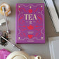 A purple book titled "Tea" is on a table surrounded by the PrintWorks Tea Tool Set, which includes a tea infuser, honey jar, stainless steel teapot, loose tea leaves, and a pink cloth.