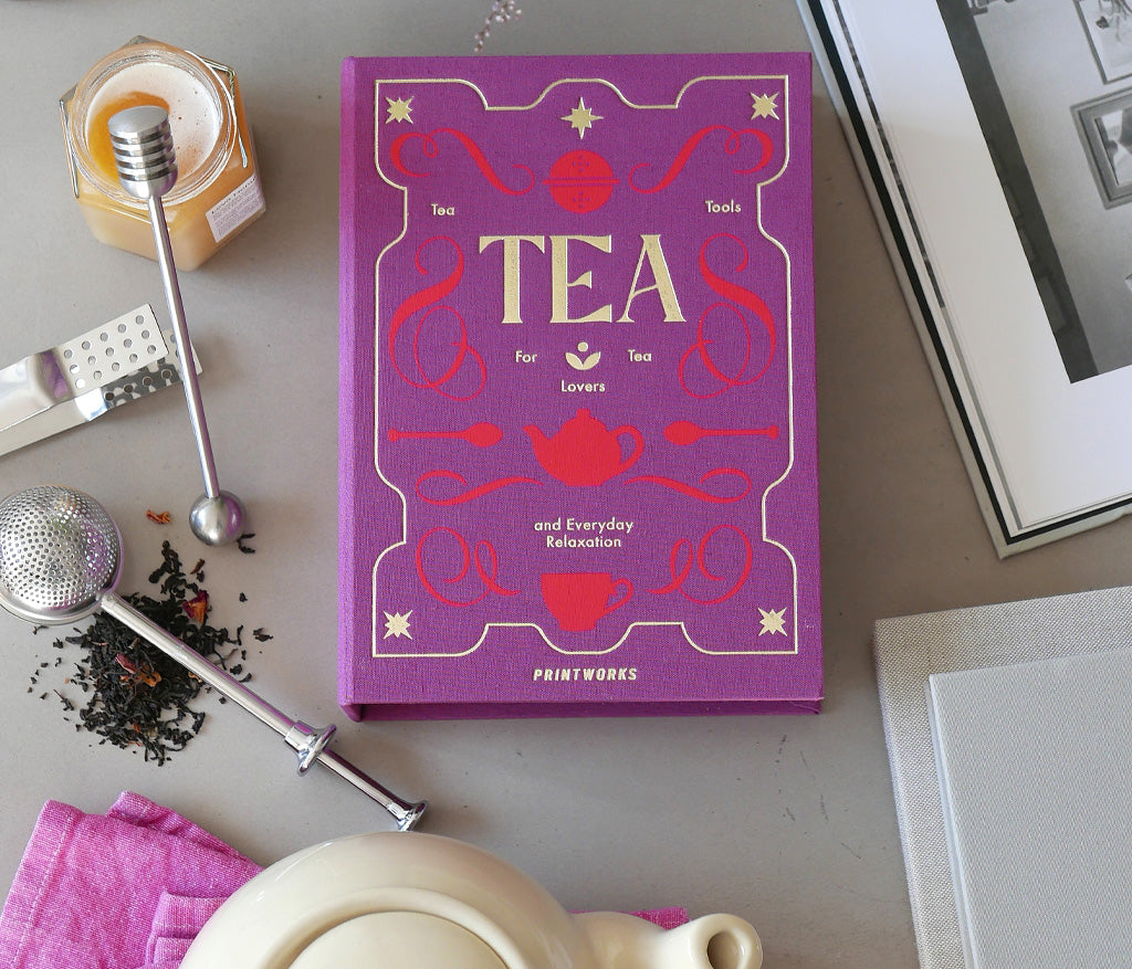 A purple book titled "Tea" is on a table surrounded by the PrintWorks Tea Tool Set, which includes a tea infuser, honey jar, stainless steel teapot, loose tea leaves, and a pink cloth.