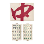 The PrintWorks Mahjong Reverra Game Set features a beige faux leather case with a bold red character on the cover, opening to reveal neatly organized mahjong tiles in two trays.