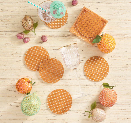 Kim Seybert's Reed Natural Coaster Set is displayed with a glass topped with a straw and umbrella, plus decorative artificial fruits, all arranged on a light wooden surface.