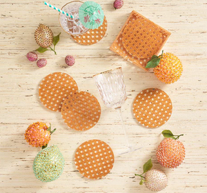 Kim Seybert's Reed Natural Coaster Set is displayed with a glass topped with a straw and umbrella, plus decorative artificial fruits, all arranged on a light wooden surface.