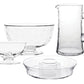 The Juliska Puro Glass Serveware Collection features a clear pitcher, large bowl, small dessert dish, and round plate with raised center—each mouth-blown with elegant textural lines and showcased on a white background.