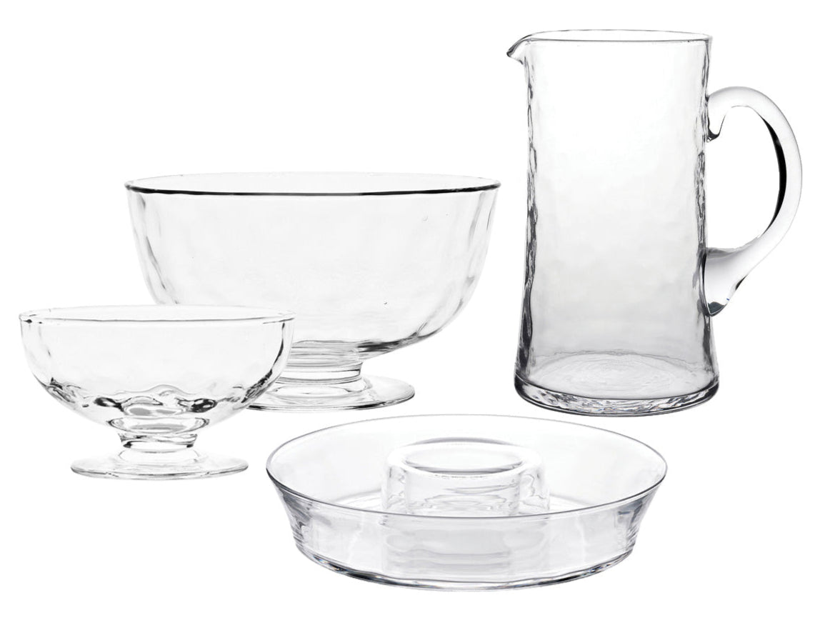 The Juliska Puro Glass Serveware Collection features a clear pitcher, large bowl, small dessert dish, and round plate with raised center—each mouth-blown with elegant textural lines and showcased on a white background.
