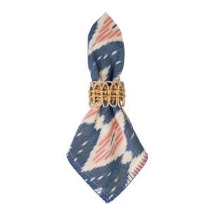 The Juliska Ikat Napkin Set features blue and white napkins with pink accents and hand-stitched trim, elegantly displayed with a woven rattan napkin ring.