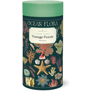 The Ocean Flora 1,000 Piece Puzzle by Cavallini Papers and Co comes in a cylindrical box with colorful ocean plant and coral illustrations on a dark green background.