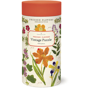 A cylindrical container labeled "Pressed Flowers 1,000 Piece Puzzle" by Cavallini Papers and Co features colorful pressed flower illustrations and an orange lid.