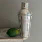 The Hôtel Silver Vintage Cocktail Shaker with Double Engine-Turned Bands, in stainless steel, stands beside a whole lime on a worn wooden surface against a gray wall, showcasing timeless Deco design.