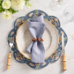 A table setting with a Kim Seybert Terraza Placemat in Blue, beige plate, folded blue napkin with wooden ring, bamboo-handled fork and knife, two glasses, and a white flower arrangement.