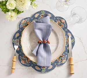 A table setting with a Kim Seybert Terraza Placemat in Blue, beige plate, folded blue napkin with wooden ring, bamboo-handled fork and knife, two glasses, and a white flower arrangement.