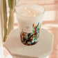 A large, multicolored Baobab Collection Rainforest Amazonia scented candle with four lit wicks on a white pedestal in a room with pink tones and a cactus.