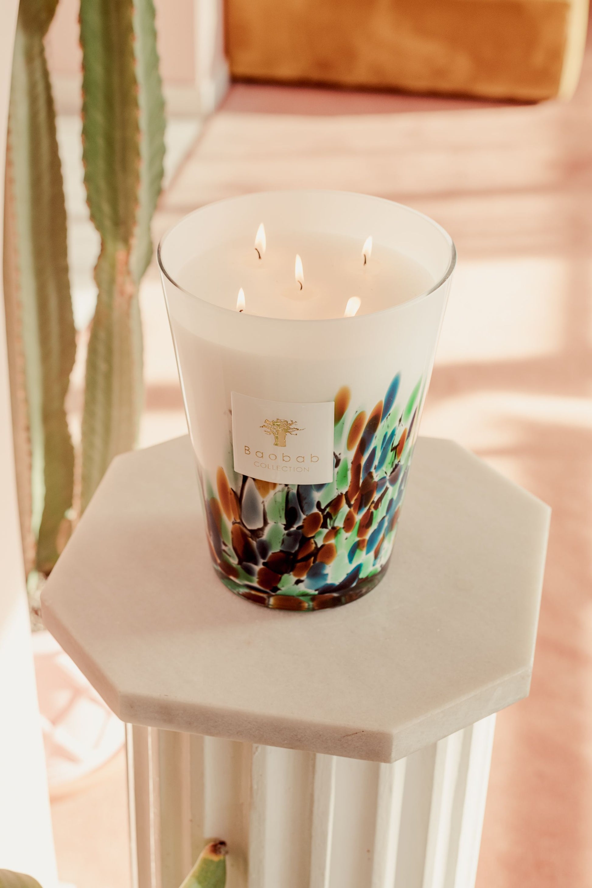 A large, multicolored Baobab Collection Rainforest Amazonia scented candle with four lit wicks on a white pedestal in a room with pink tones and a cactus.
